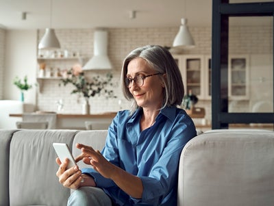 The picture shows a woman sitting peacefully on her couch and looking at her phone while smiling. background of the picture ist slightly blurred but you can still distinguish is a white modern kitchen with a white stone wall. On the stone wall are two simple shelves with food containers and to the right of them on the wall is an extractor bonnet. On the right-hand side of the picture, three wall cabinets can be seen, two of which have glass fronts. Underneath you can see a counter with two white chairs. On the counter is a vase with decorative flowers. In the foreground you can see the upper part of a light grey couch on which an elderly lady is sitting. She has a grey bob, black glasses and is wearing a blue shirt and light blue jeans. The lady is smiling at her mobile phone she is holding in her hand, with which she can easily and conveniently use ista's SmartPay option.