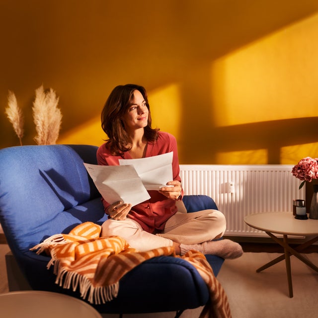 Eine brünette Frau sitzt im Schneidersitze auf ihrer blauen Couch im Wohnzimmer. In den Händen hält Sie zwei Seiten Papier, Seiten aus der Heizkostenabrechnung von ista. Sie blickt zufrieden Richtung Fenster. 