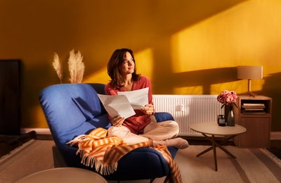 Eine brünette Frau sitzt im Schneidersitze auf ihrer blauen Couch im Wohnzimmer. In den Händen hält Sie zwei Seiten Papier, Seiten aus der Heizkostenabrechnung von ista. Sie blickt zufrieden Richtung Fenster. 