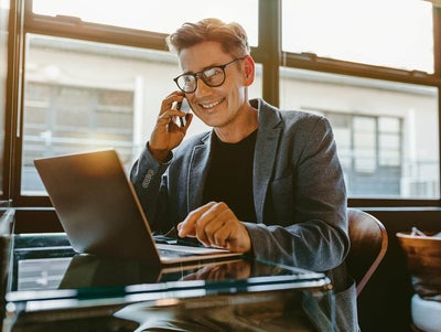 The picture shows a man who is apparently sitting in a café or restaurant with his laptop at a single seat with a glass table. He is sitting at the edge, right next to a window front that can be seen in the background. He is wearing a grey flowing jacket and a simple black jumper underneath. He has short brown hair and wears square black glasses. He is holding his mobile phone to his ear with his right hand and appears to be making a call as he smiles broadly. His left hand is placed on the touchpad of his laptop.