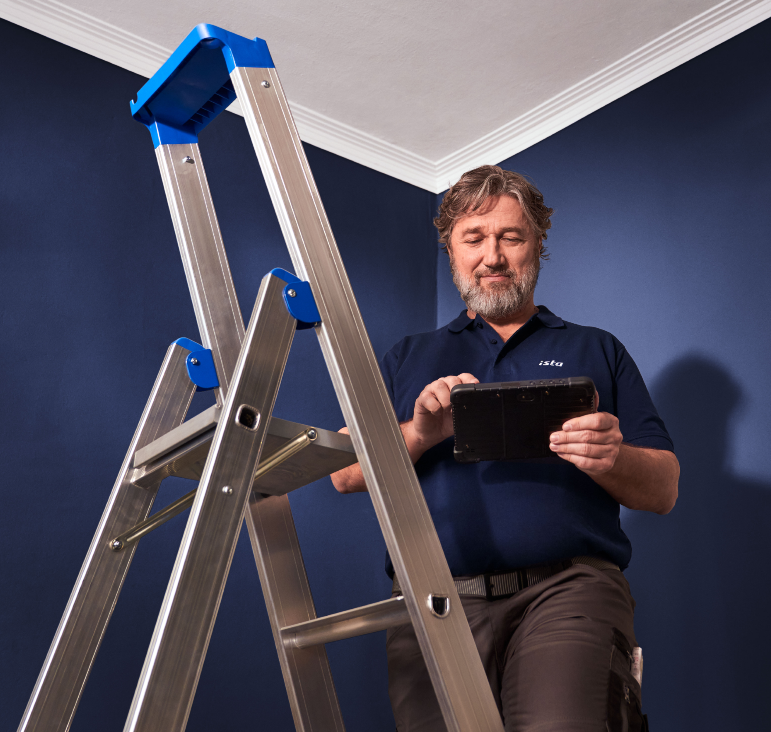 Ein ista ServicePartner steht neben einer silbernen Klappleiter vor einer dunkelblauen Wand und blickt auf ein Tablet in seinen Händen. Er trägt ein dunkelblaues T-Shirt mit weißem ista Logo und eine braune Hose mit Gürtel.